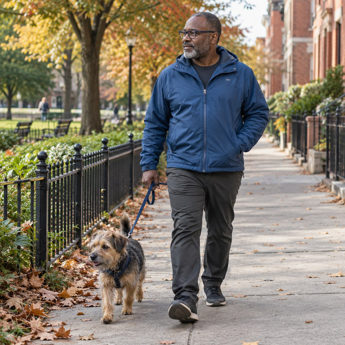Man walking a small terrier beside a neighborhood park in autumn